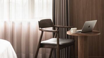Una habitación de hotel con una silla de madera, una mesita redonda pequeña con una computadora portátil y una taza blanca, una cama y una ventana con cortinas de encaje.