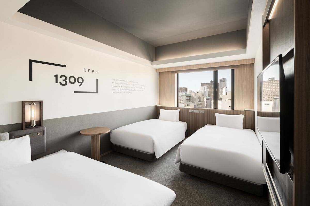 A hotel room with two beds, a small table, a wall-mounted TV, and a city view.