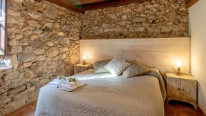 A bedroom with a stone wall, a wooden headboard, a bed with a textured blanket, two bedside tables with lamps, and a window with a view.