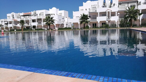 A swimming pool with a clear blue water surface, surrounded by white buildings with balconies and palm trees.