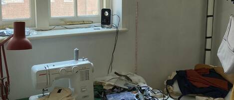 A sewing room with a sewing machine, a window with a view of brick buildings, a lamp, and various sewing materials on a table.