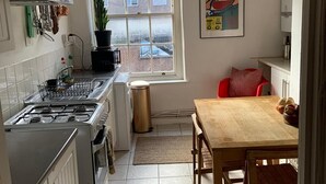 A compact kitchen with a gas stove, white cabinets, a window with a view, a dining table with chairs, and a hanging light fixture. - Charming 3-Bed Apartment in London (London)