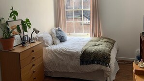 A bedroom with a bed, wooden dresser, potted plant, and a window with curtains. - Charming 3-Bed Apartment in London (London)