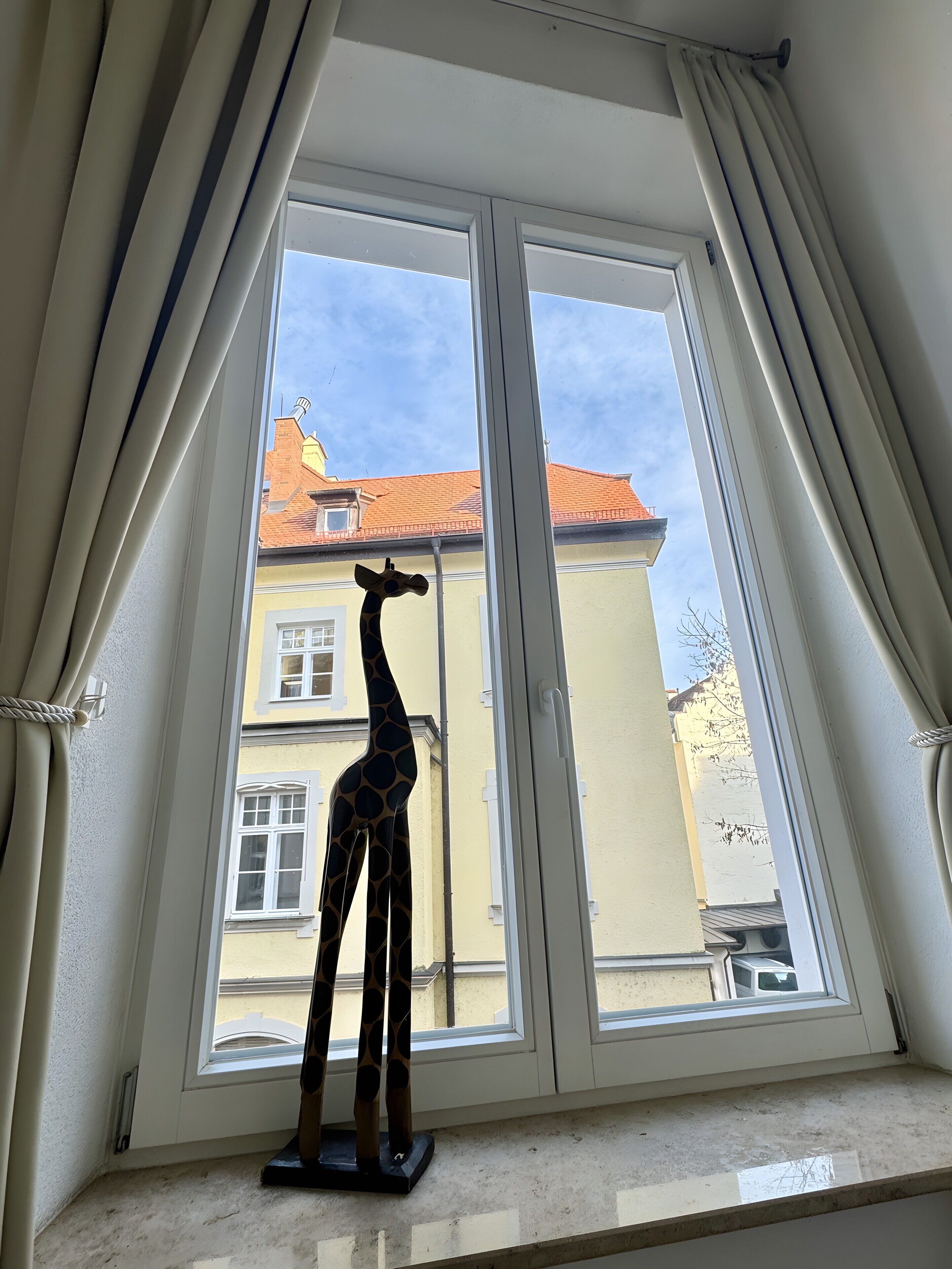 A room with a large window, a giraffe sculpture, and a view of buildings outside.