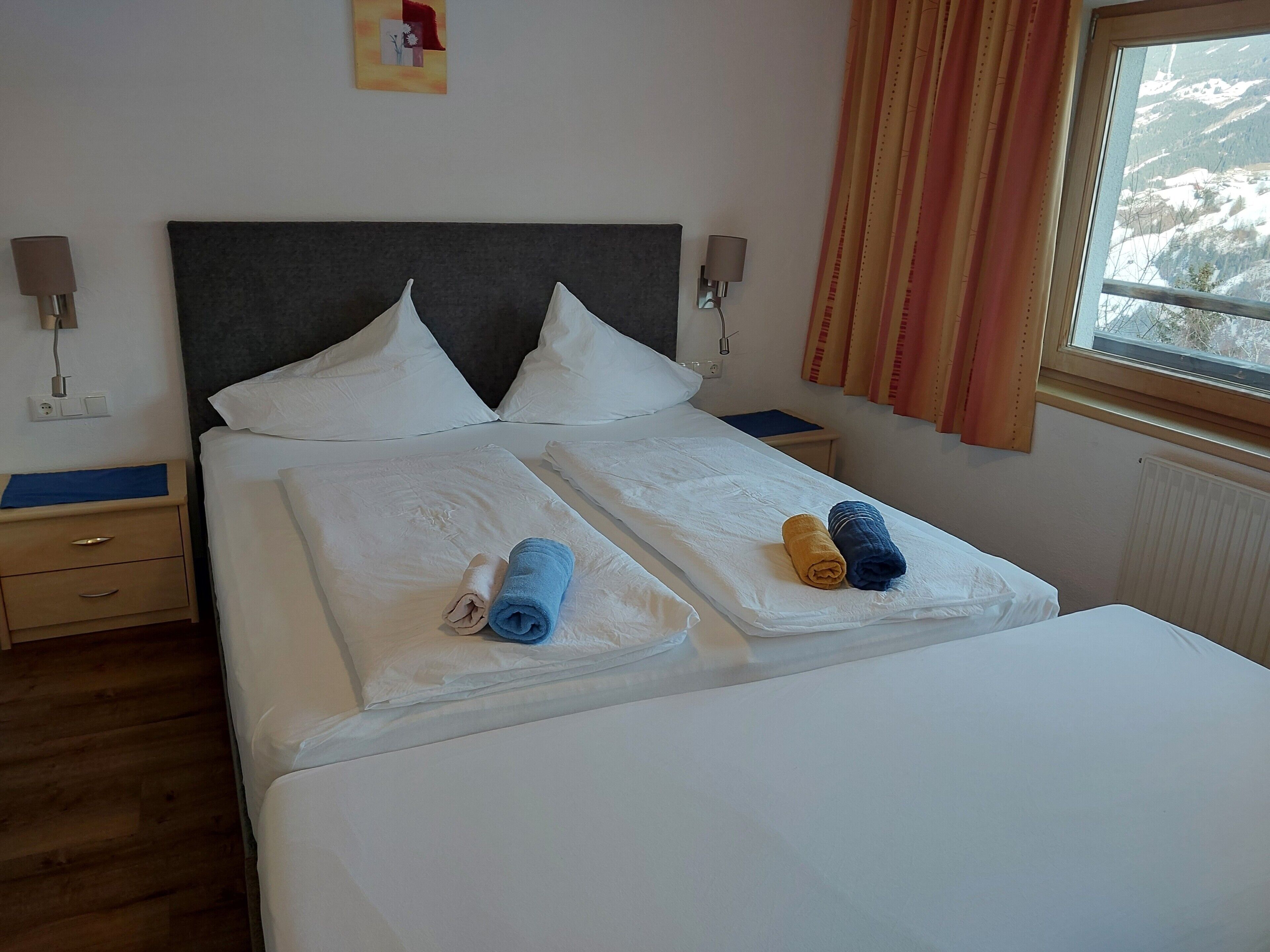 Cama bien hecha con sábanas blancas, dos almohadas y dos toallas enrolladas en azul y amarillo.
