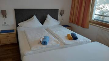 Cama bien hecha con sábanas blancas, dos almohadas y dos toallas enrolladas en azul y amarillo.