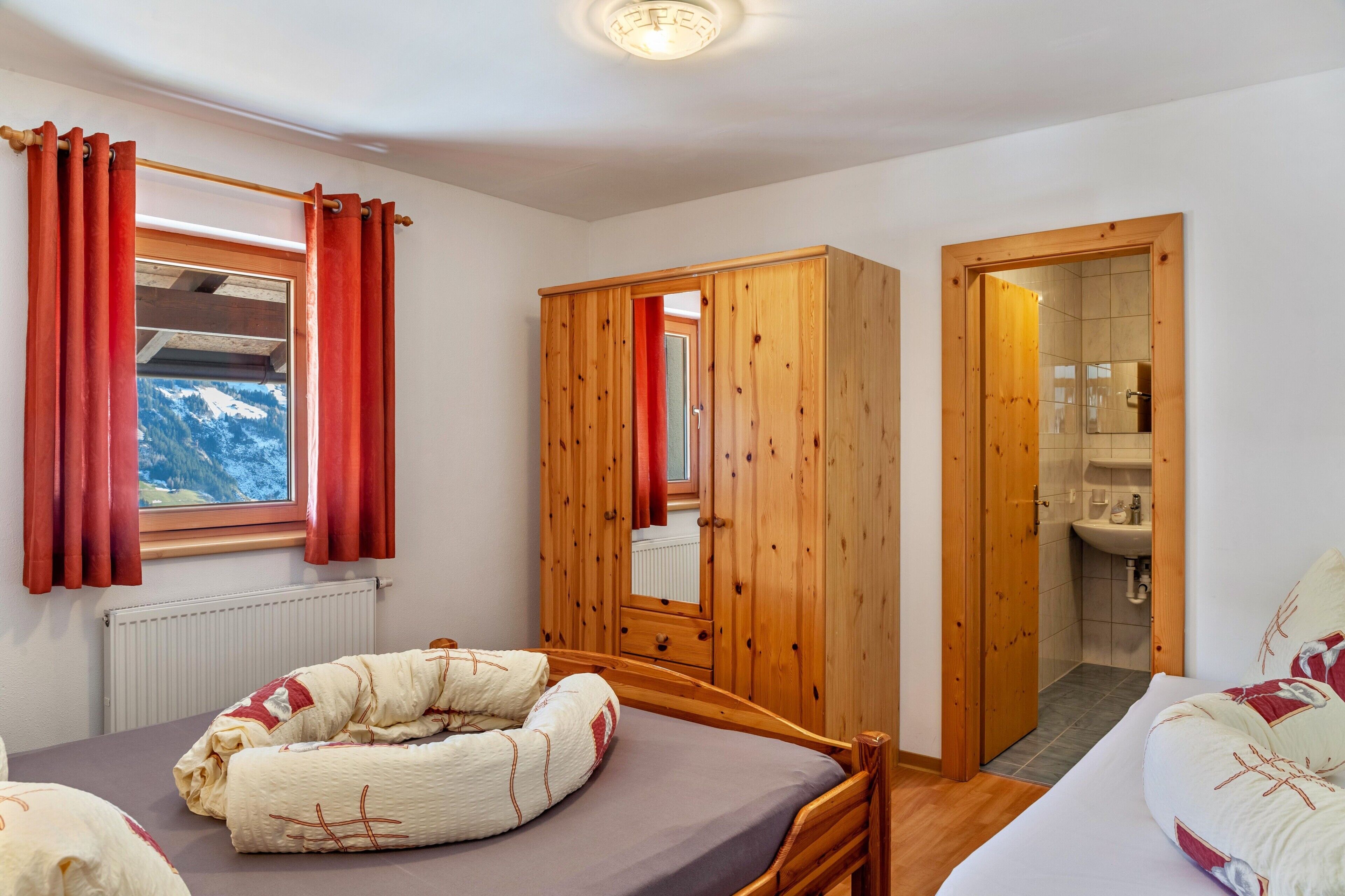 A bedroom with a bed, a wooden wardrobe, a bathroom with a sink, and a window with curtains.