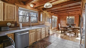 L’intérieur d’une cabane en bois comprend une cuisine et un coin repas, avec des meubles de rangement en bois, un réfrigérateur en acier inoxydable et une grande fenêtre qui laisse entrer la lumière naturelle.