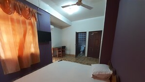 Une chambre avec un lit, un ventilateur de plafond, une télévision et une salle de bain visible à travers une porte ouverte.