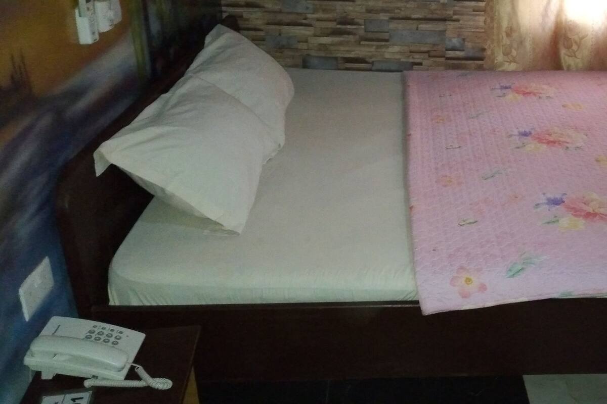 A single bed with a white pillow and a floral blanket, a bedside telephone, and a stone wall behind the bed.