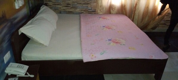 A single bed with a white pillow and a floral blanket, a bedside telephone, and a stone wall behind the bed.