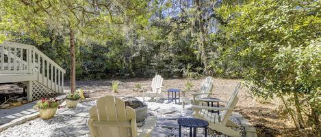 A patio area with Adirondack chairs, a small table, and a bench, surrounded by white pebbles and greenery.