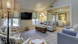 A spacious living room with a high ceiling, a large window, a flat-screen TV, wicker furniture, and a painting on the wall. - Sunset Rentals | Lawton 45 | Dog Friendly | (Hilton Head Island)