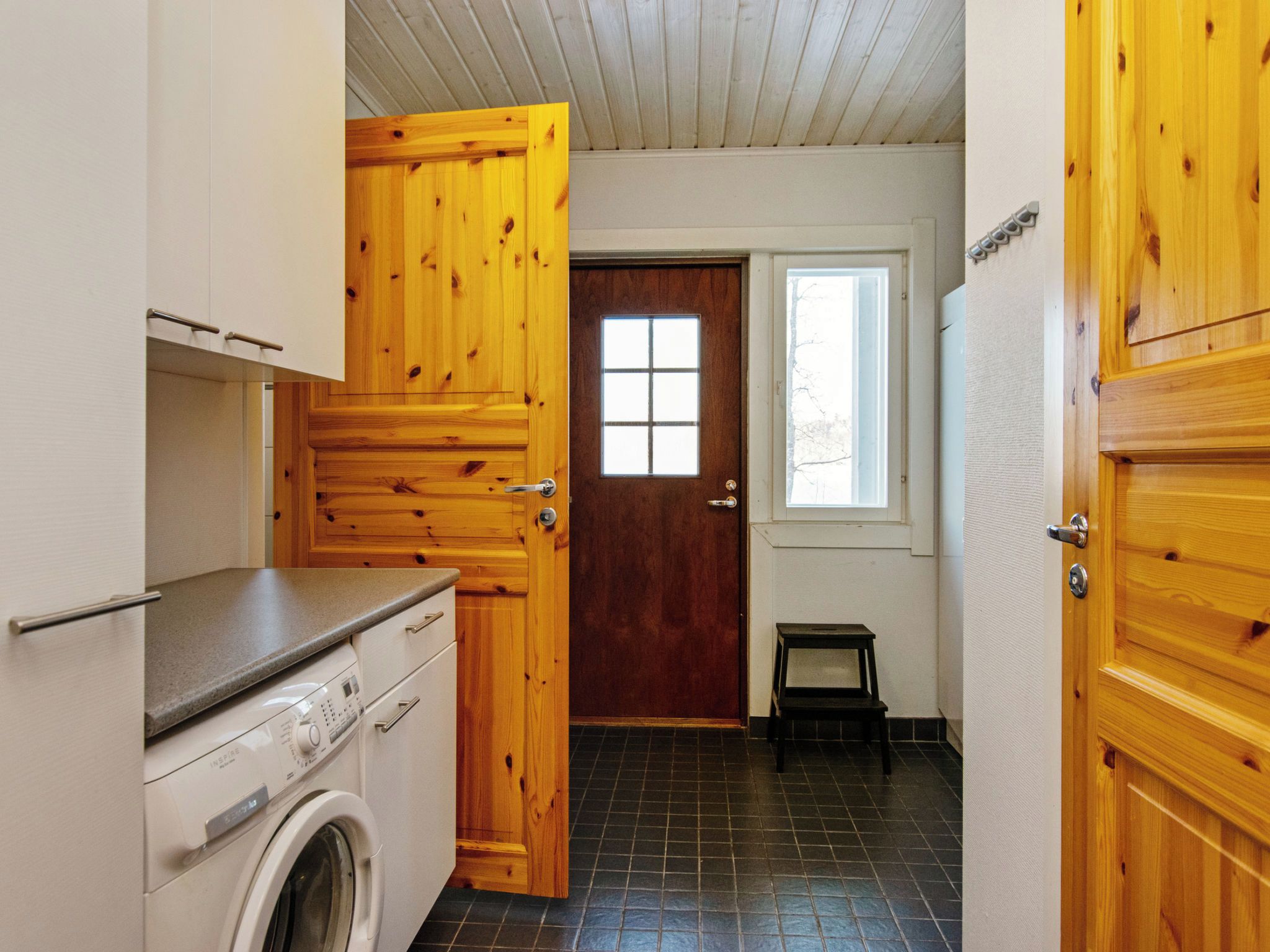 A laundry room with a washing machine, wooden cabinets, a small stool, and a door with a window.