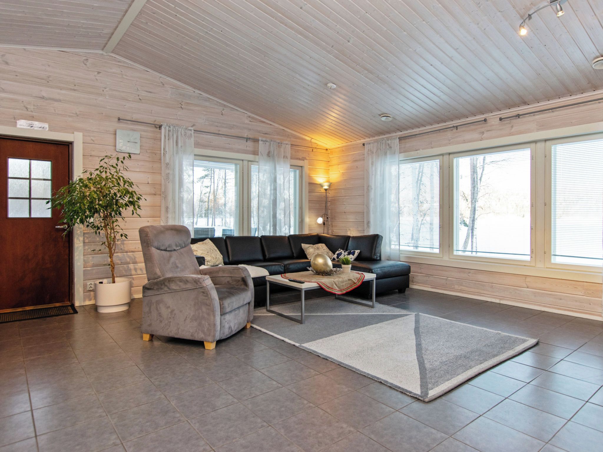 A spacious living room with a sofa, armchair, and coffee table, large windows, and a wooden ceiling.