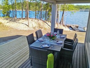 Un espace de restauration en plein air avec une table dressée pour quatre personnes, offrant une vue sur une étendue d’eau et une zone boisée.