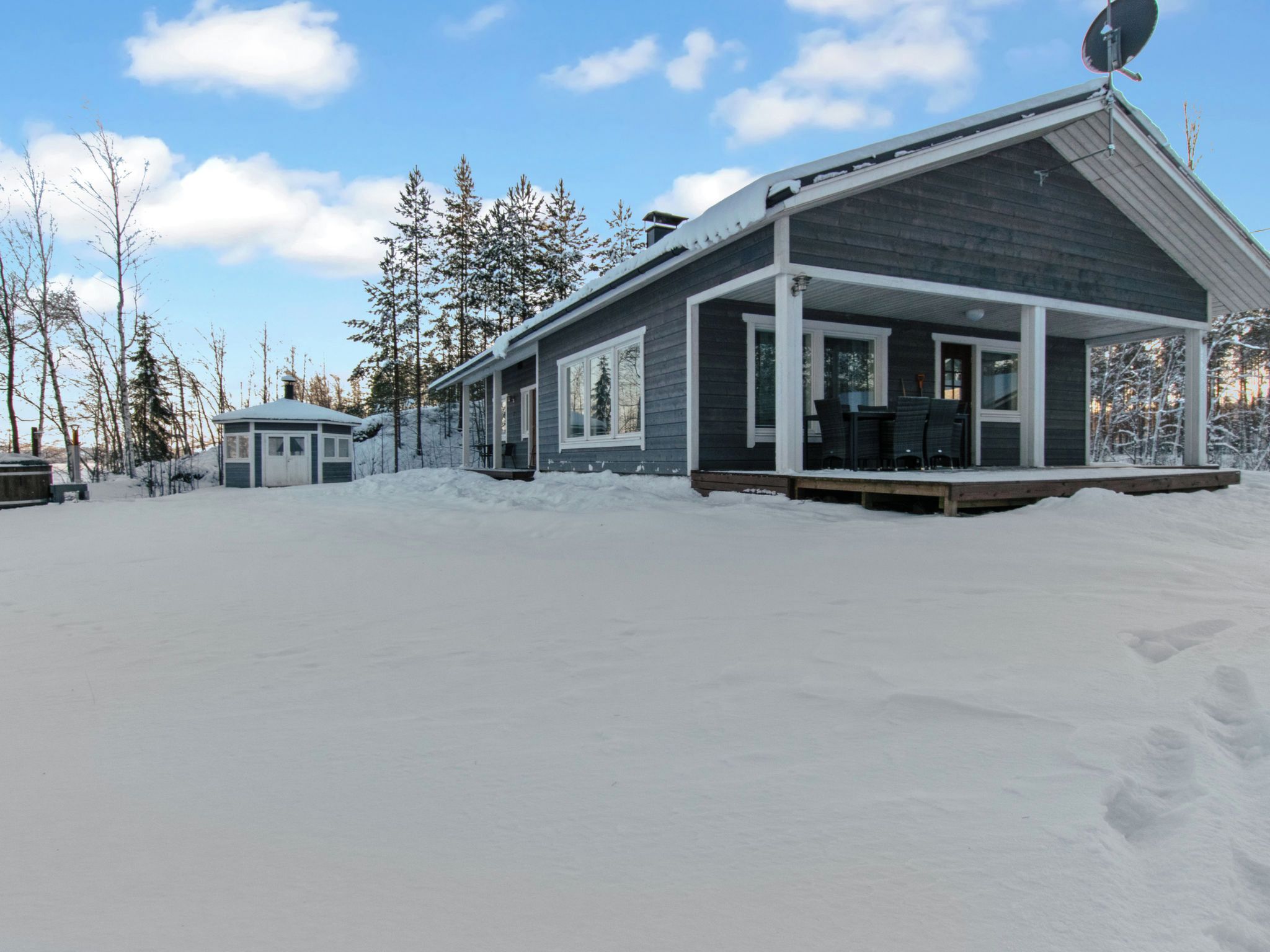 Una cabaña moderna con un porche cubierto y una antena parabólica, rodeada de terreno y árboles cubiertos de nieve.