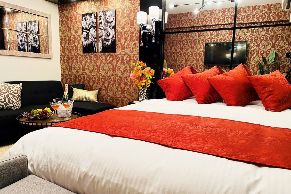 Une chambre d’hôtel avec un grand lit, des oreillers rouges, un canapé noir, une petite table avec des boissons et des fruits, et des œuvres d’art accrochées aux murs.
