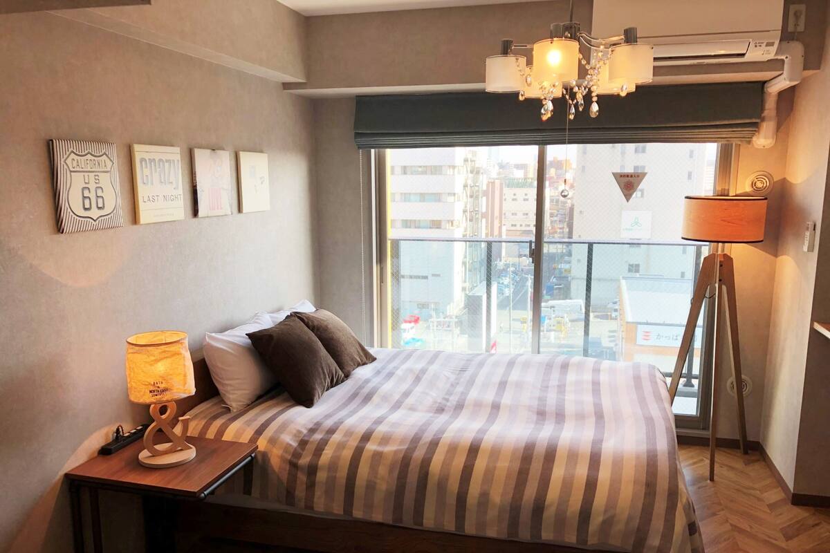 Une chambre d’hôtel comprenant un lit, une table de chevet avec une lampe, une fenêtre donnant sur des immeubles et des éléments de décoration murale, notamment un panneau de signalisation routière et des tableaux encadrés.