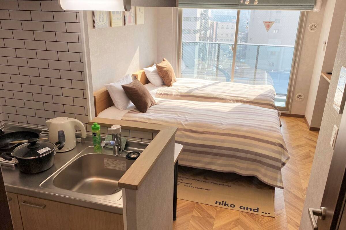 Une chambre d’hôtel compacte, dotée d’une kitchenette, d’un lit avec une literie à rayures et offrant une vue sur la ville par la fenêtre.