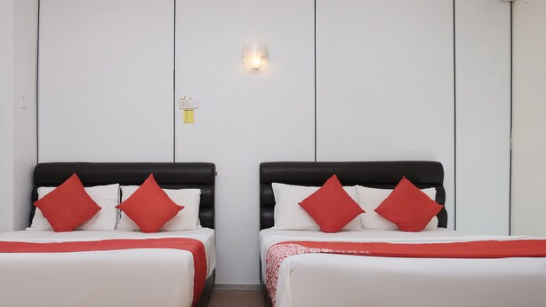Duas camas com cabeceiras pretas e almofadas vermelhas, roupa de cama branca e uma pequena luminária fixada na parede.