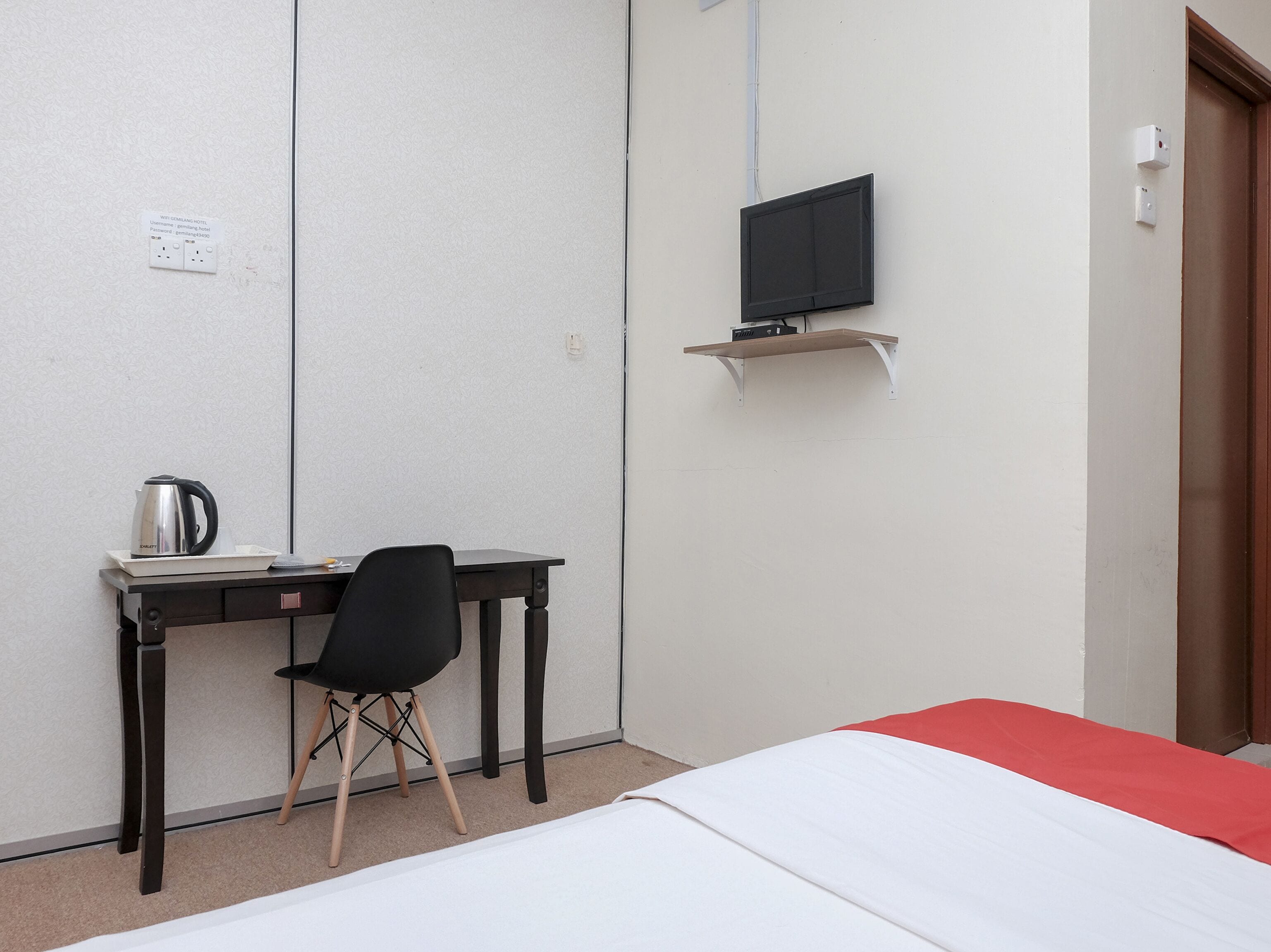 Een hotelkamer met een bed, een bureau met stoel, een televisie op een plank en een waterkoker op het bureau.