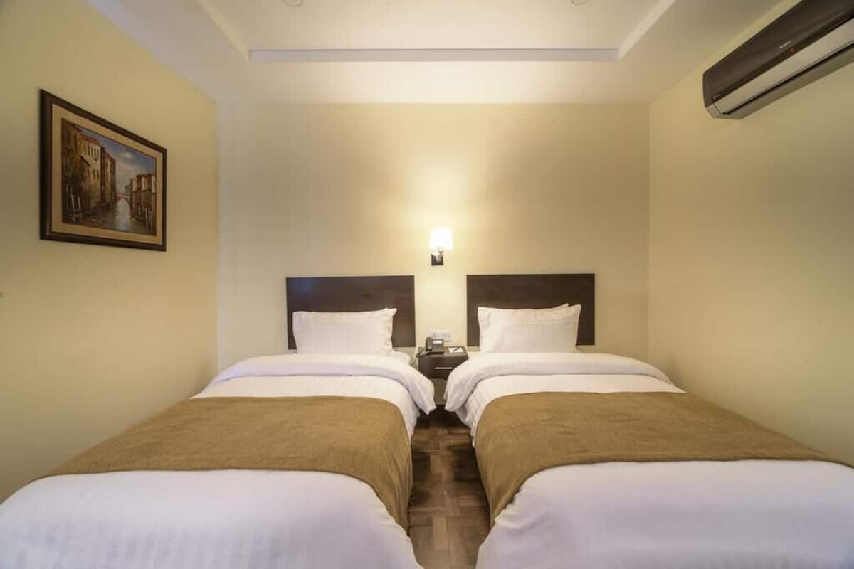 A hotel room with two beds, a nightstand between them, a wall-mounted lamp, and a painting of a canal scene.