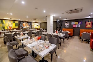 A restaurant with multiple dining tables and chairs, a bar area, and a wall with various artworks. - Hotel One Sahiwal (Sahiwal)