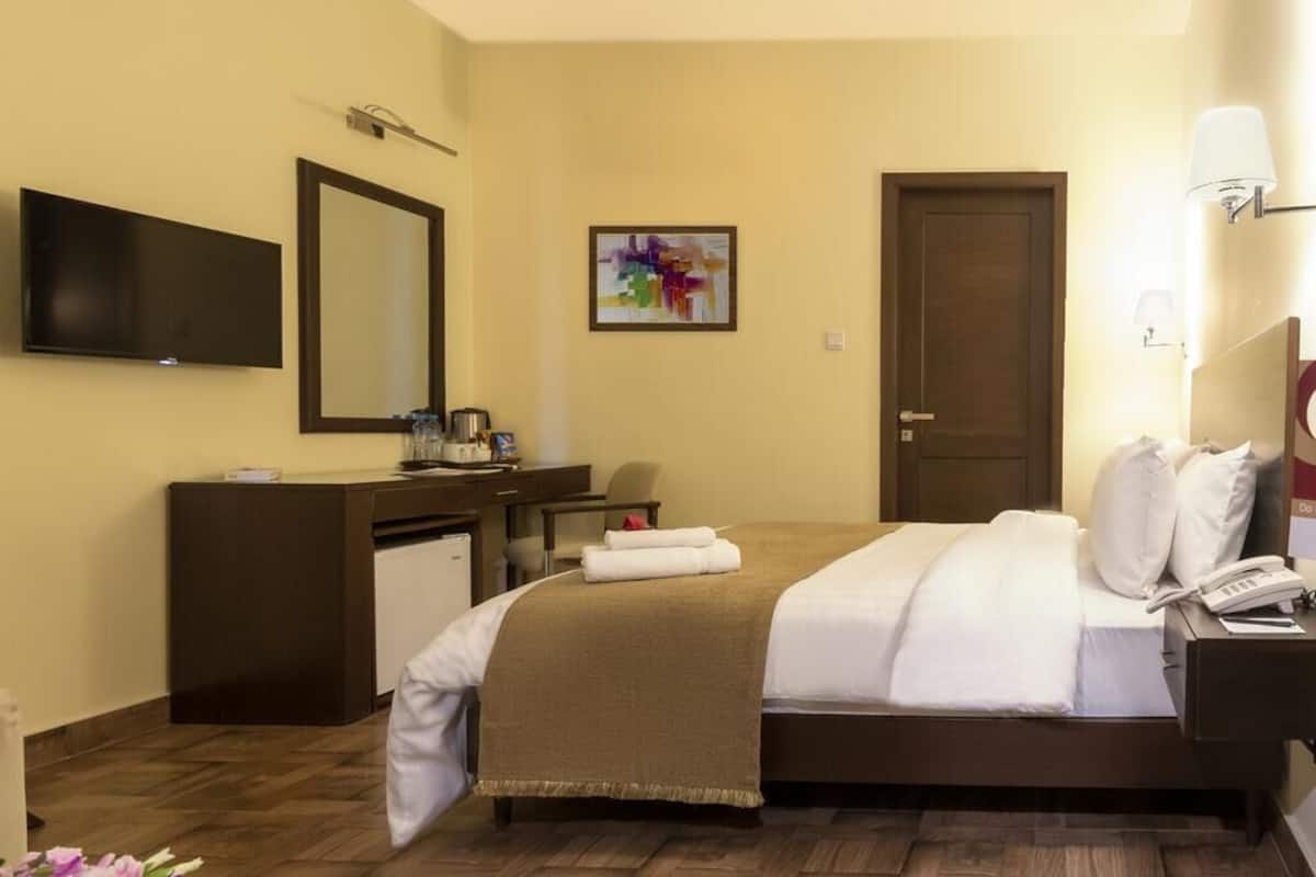 A hotel room with a bed, a desk with a chair, a television, a door, and a painting on the wall.
