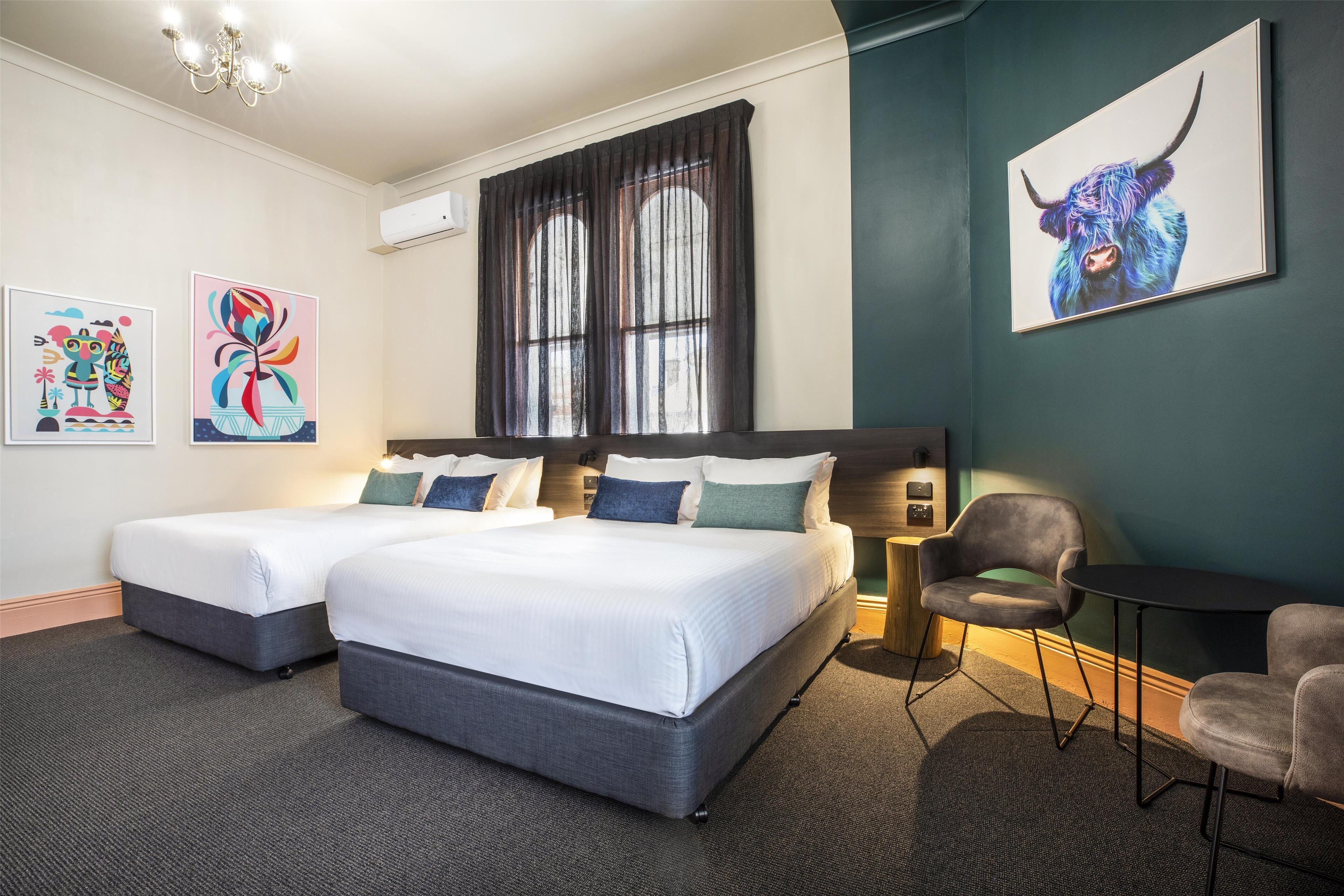 A hotel room with two beds, a desk, and chairs. There are two paintings on the wall and a window with curtains.