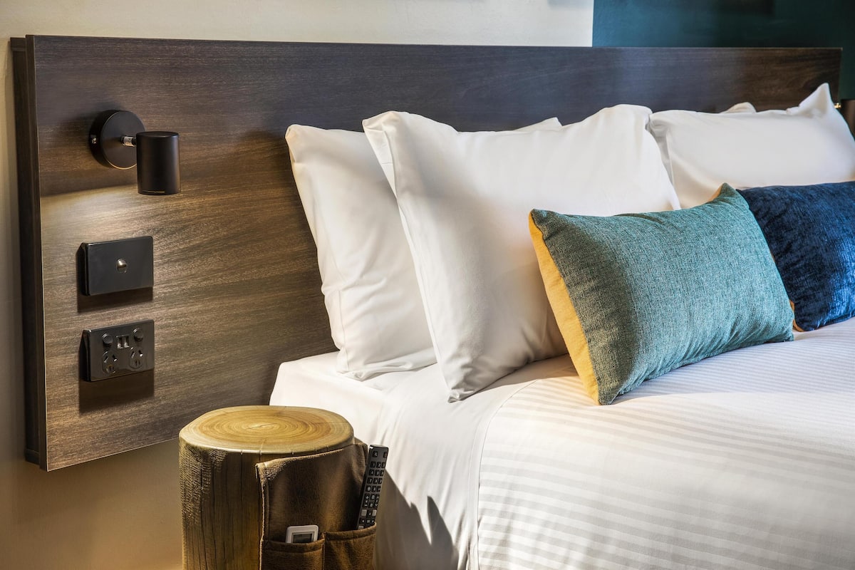 A neatly made bed with white and teal pillows, a wooden headboard, and a bedside table with a remote control.