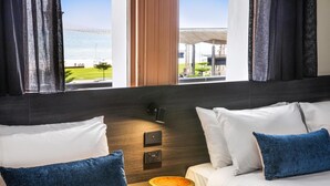 A hotel room with two beds, a wooden nightstand, a wall-mounted control panel, and a view of a modern building and greenery outside. - Ramsgate Hotel by Nightcap Social (Henley Beach)