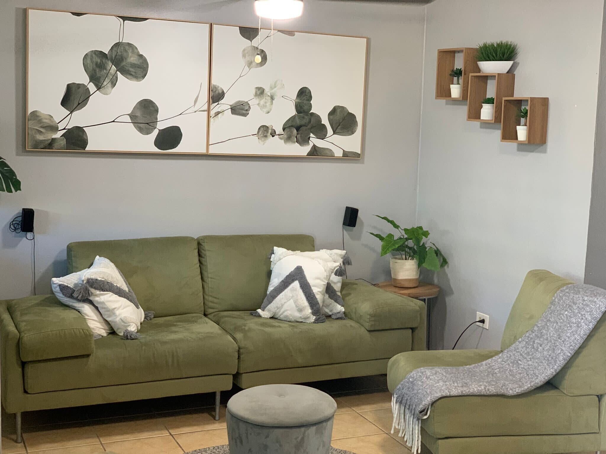 A green sofa with two pillows, a grey ottoman, and a grey throw blanket. There is a potted plant on a side table.