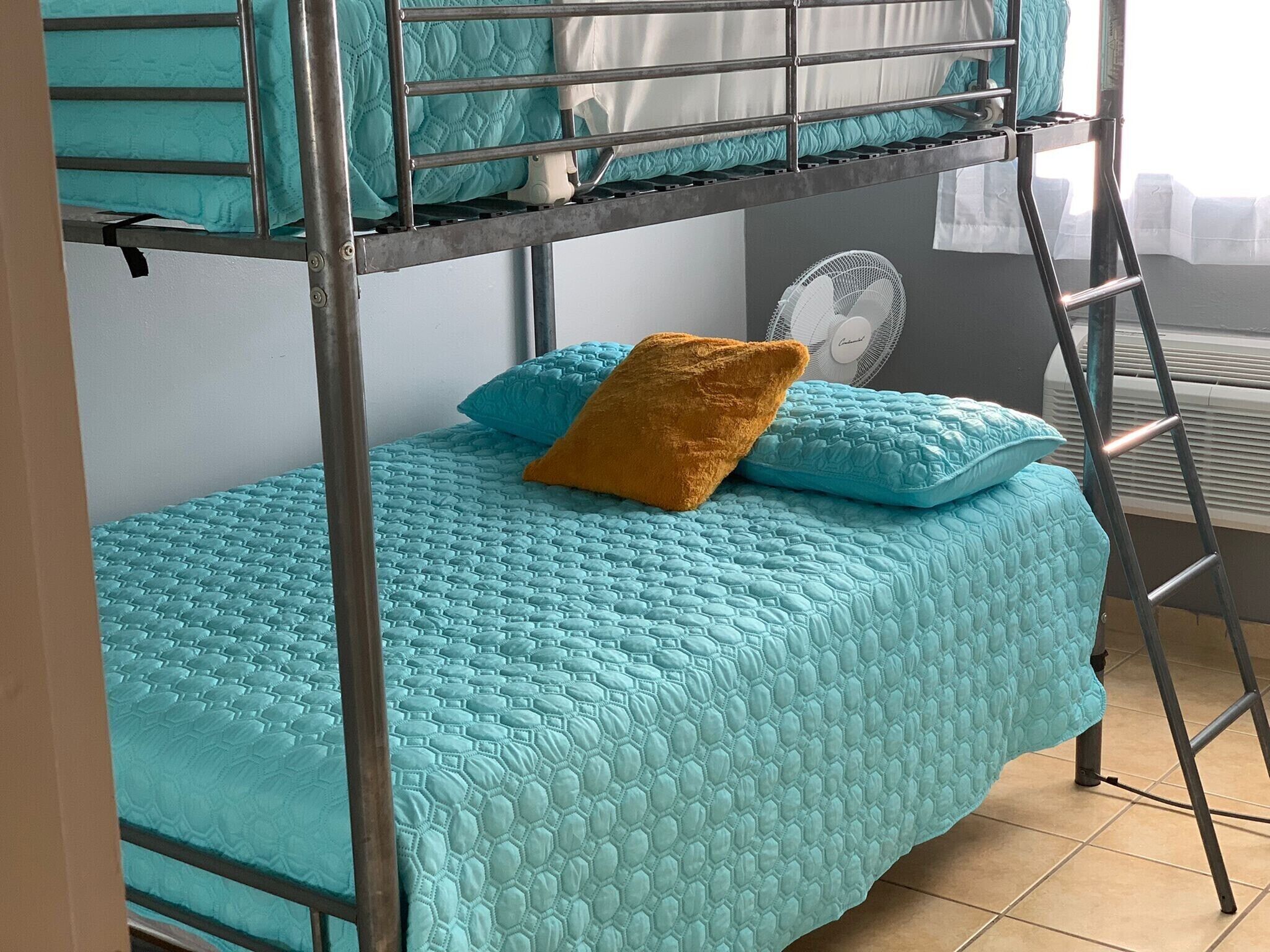 Un letto a castello con materasso e cuscini trapuntati color acquamarina, una ventola e una scala.