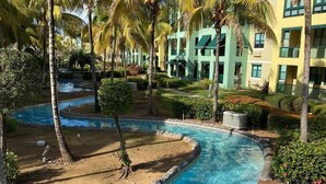 A pool area with a clear blue pool, surrounded by palm trees and greenery, and adjacent to a building with multiple windows and balconies. - Aquatika “A Cozy Paradise” (LOIZA)