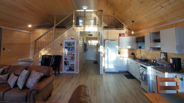 Un amplio salón con escalera de madera, una cocina con armarios blancos y una estantería.