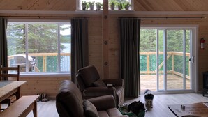 Interior acogedor de una cabaña con techo de madera, un sofá, una mesa de comedor y un ventanal con vistas a una terraza y al agua.