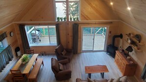 Una habitación acogedora estilo loft, con techos altos, paredes de madera, un amplio ventanal arqueado, un ventilador de techo, chimenea y zona de comedor con banco y sillas.