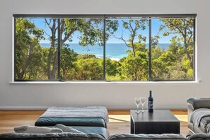 A room with a large window offering a view of the sea and trees, a bed, a coffee table with glasses and a bottle, and a sofa.