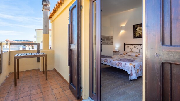 A balcony with a wooden table, an open door leading to a bedroom with a bed and a nightstand, and a wall-mounted light fixture.