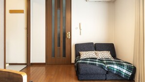 A room with a wooden floor, a dark sofa with patterned cushions, a wooden door with glass panels, and a wall-mounted air conditioner. - Urban Stay Shinagawa (Tokyo)
