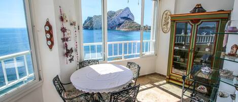 A room with a round table and chairs, a ceiling fan, a cabinet with glass doors, and a view of the sea through large windows.