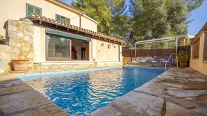 A poolside area with a stone pool, a covered patio with chairs, and a house with a stone facade and green shutters.