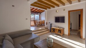 A modern living room with a grey sofa, wooden beams on the ceiling, a TV mounted on the wall, and a dining area with wooden furniture.