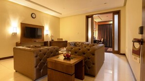 A modern hotel room with a large L-shaped sectional sofa, a wooden coffee table, a flat-screen TV mounted on the wall, and a dining area visible through an open door. - Hotel One Tariq Road Multan (Multan)