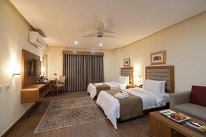 A hotel room with two beds, a desk, a chair, a TV, an air conditioner, a ceiling fan, and a small table with fruit. - Hotel One Tariq Road Multan (Multan)