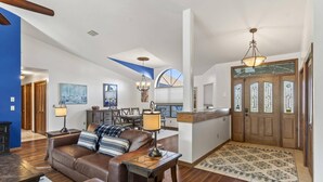 A spacious living room with a brown leather sofa, a wooden coffee table, and a dining area with a wooden table and chairs. There is a large window providing natural light and a view of the outdoors.