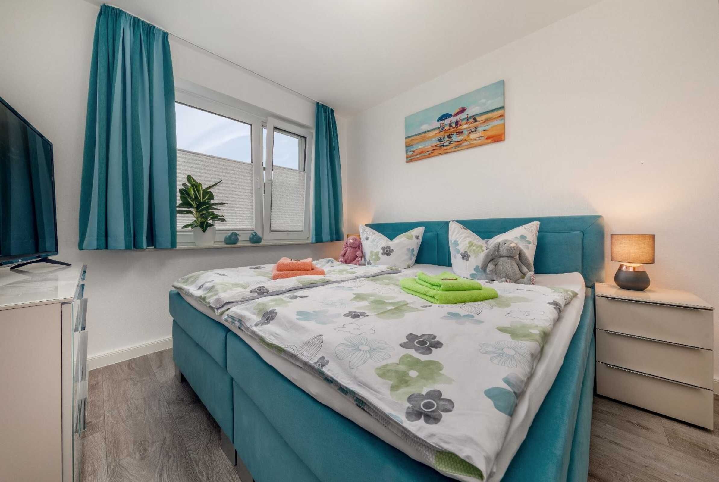 Une chambre à coucher avec un lit, une fenêtre avec des rideaux, une table de chevet avec une lampe, un tableau au mur et une plante en pot.