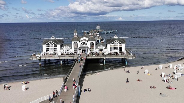 Une jetée s’avançant dans la mer, avec un bâtiment à son extrémité, une plage de sable et des personnes qui se promènent.