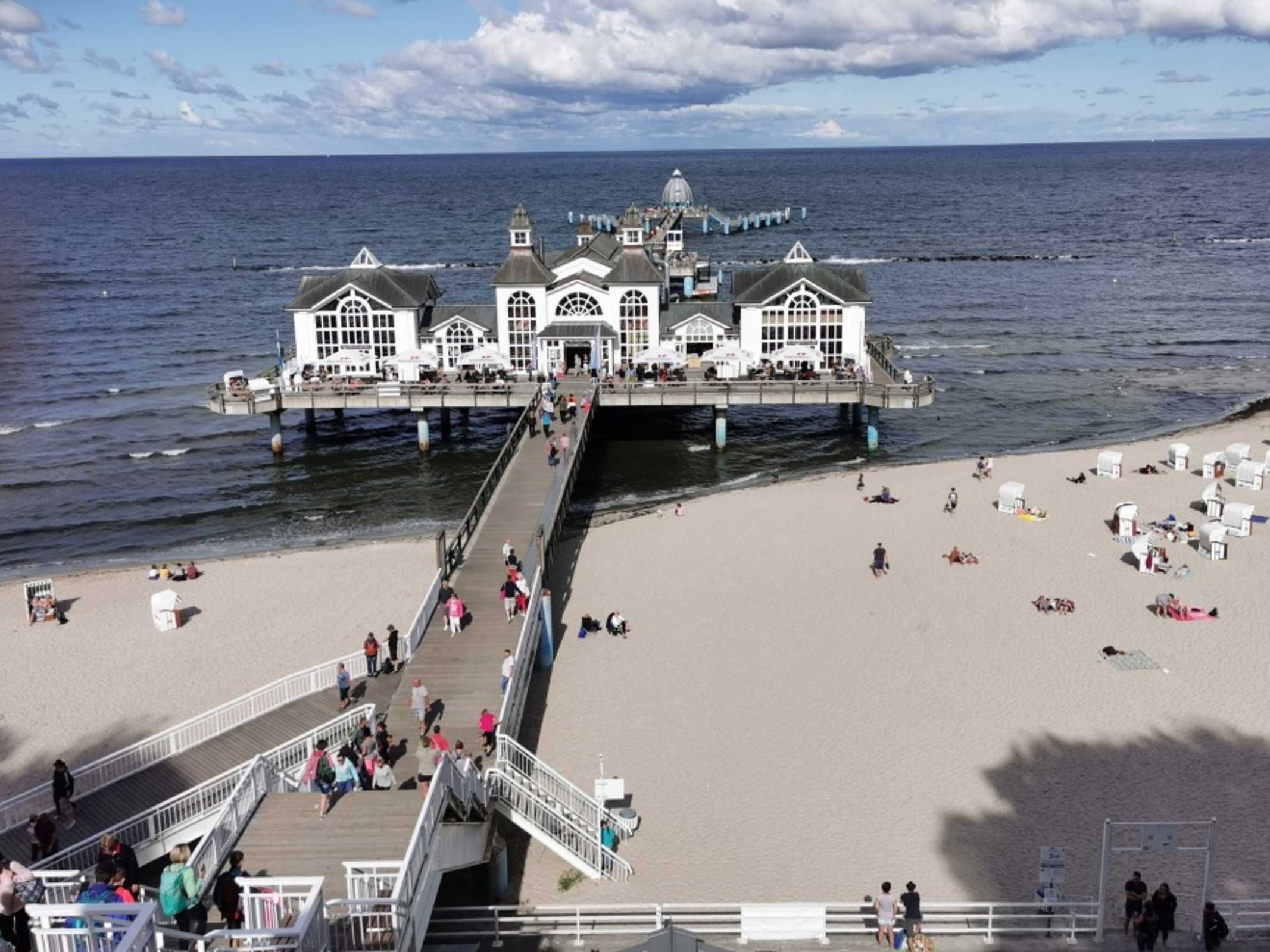 Un moll que s'endinsxa a la mar amb un edifici al final, una platja de sorra i persones passejant.
